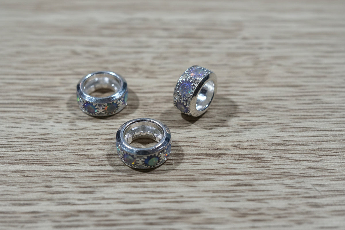 Three silver-toned spacer bead with multicolor iridescent glitter, arranged on a light brown wood grain surface.