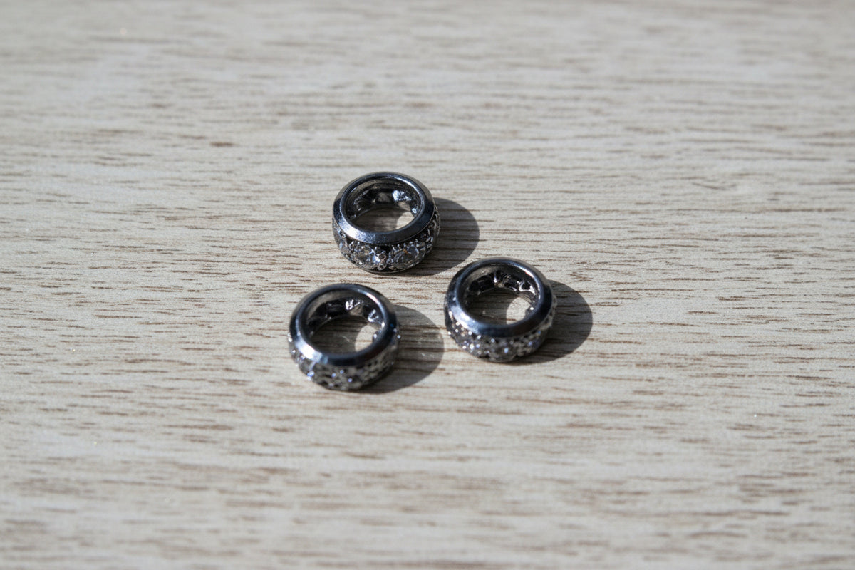metallic spacer beads with a textured, hammered finish and clear rhinestone-like embellishments, arranged on a light brown wood grain surface.