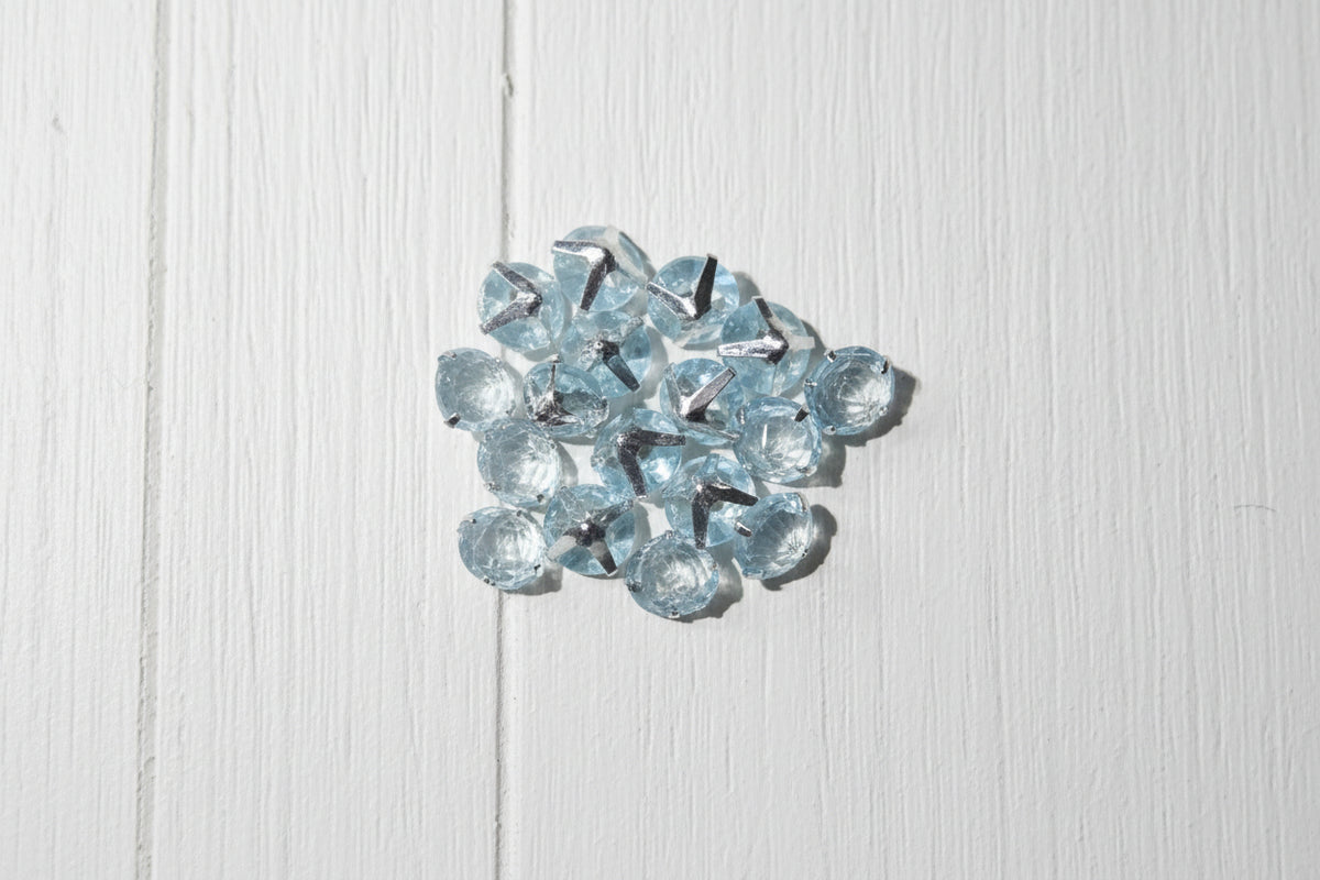A group of small, faceted light blue zircon beads scattered on a textured wood grain surface.