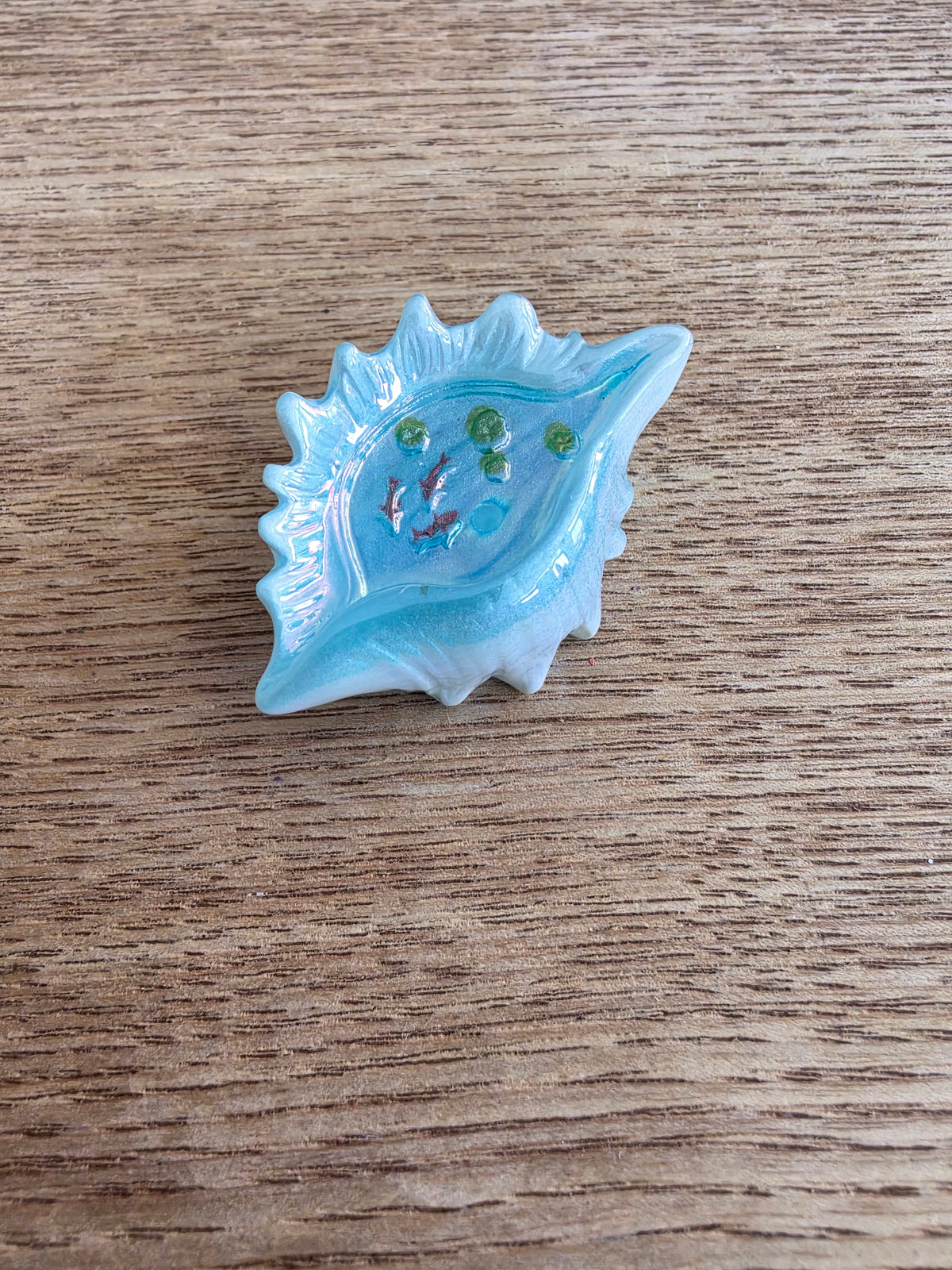 A small light blue seashell-shaped dish or tray with a glossy finish, containing tiny decorative fish and green beads, placed on a brown wood-grain surface.