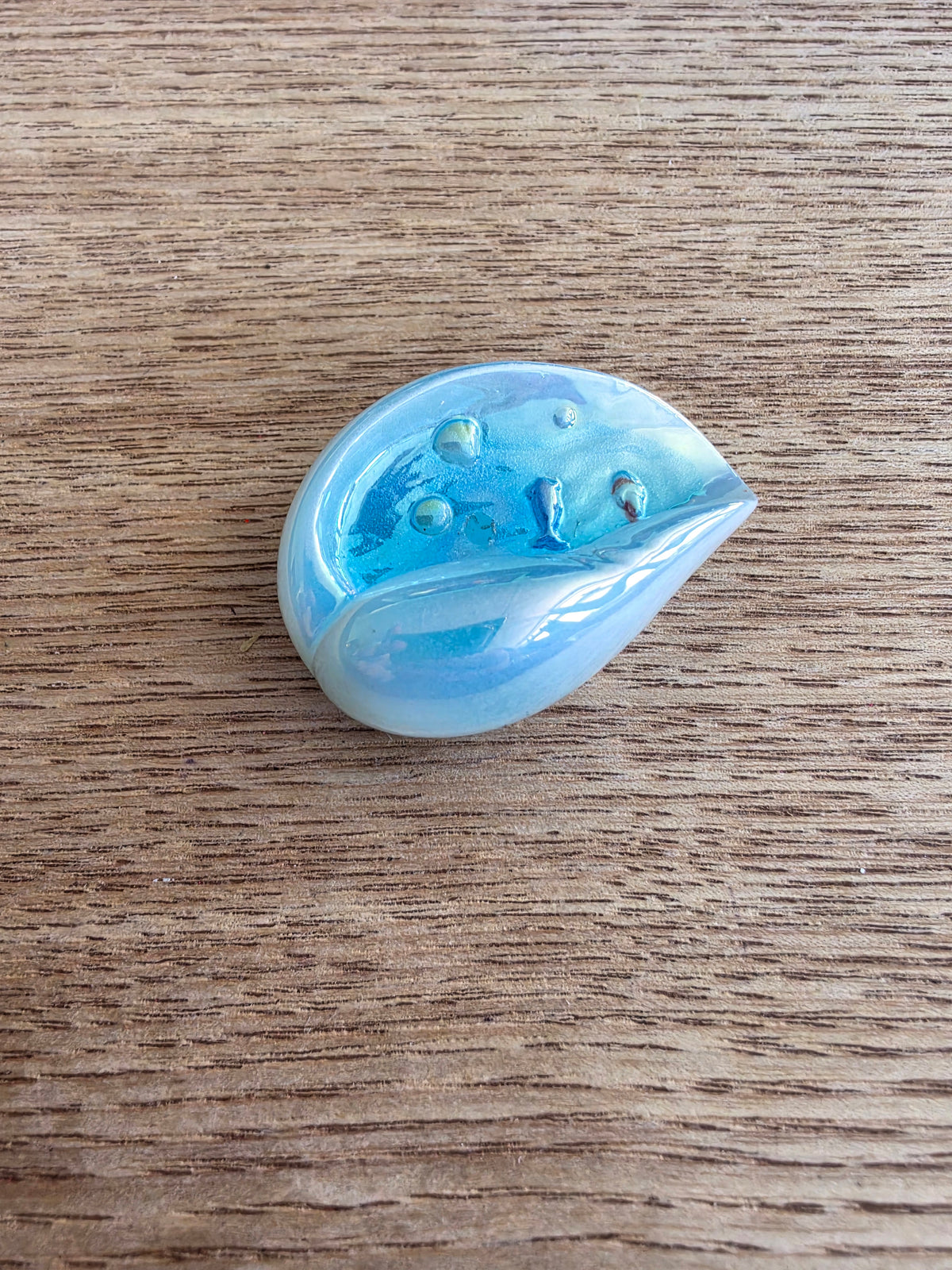 A small teardrop-shaped resin bead in iridescent light blue and white, resting on a brown wood-grain surface.