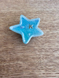 A small five-pointed star-shaped blue bead with a textured, slightly glossy finish, resting on a brown wooden surface.