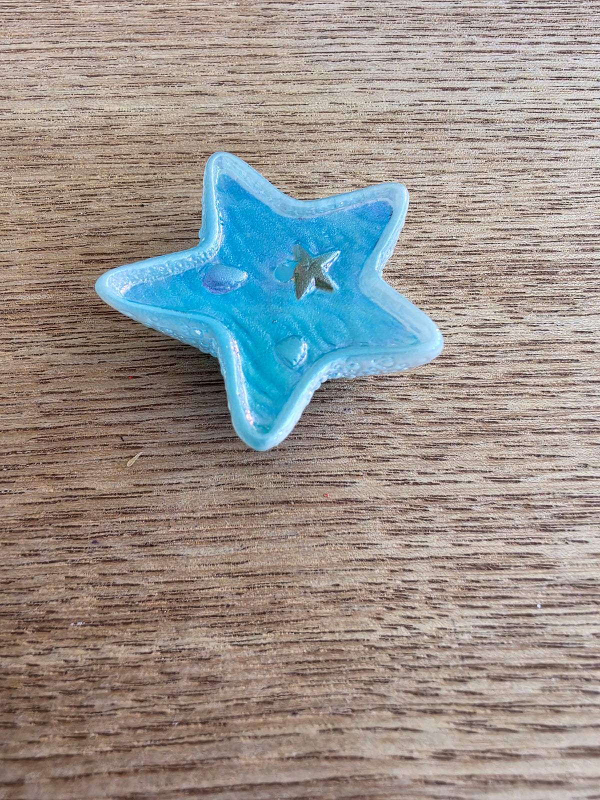 A small five-pointed star-shaped blue bead with a textured, slightly glossy finish, resting on a brown wooden surface.