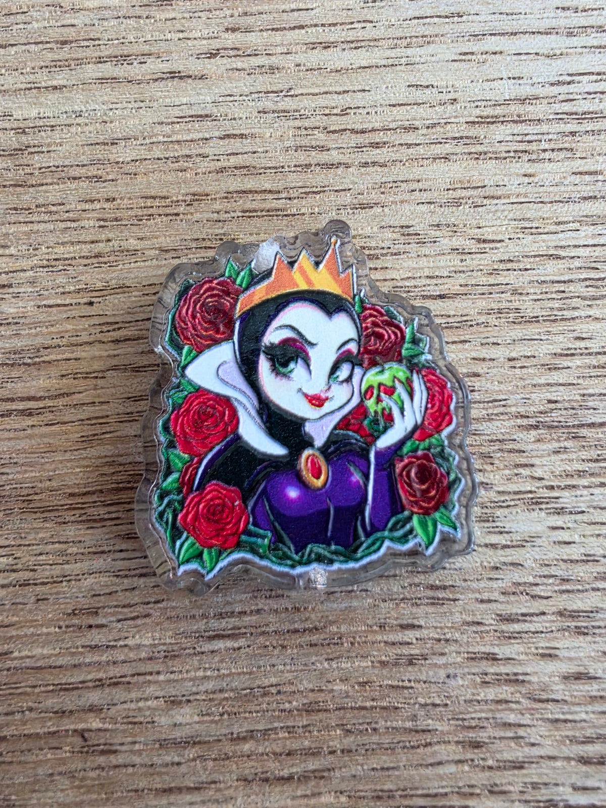 An acrylic bead featuring a stylized cartoon queen surrounded by red roses, placed on a light wood grain surface.