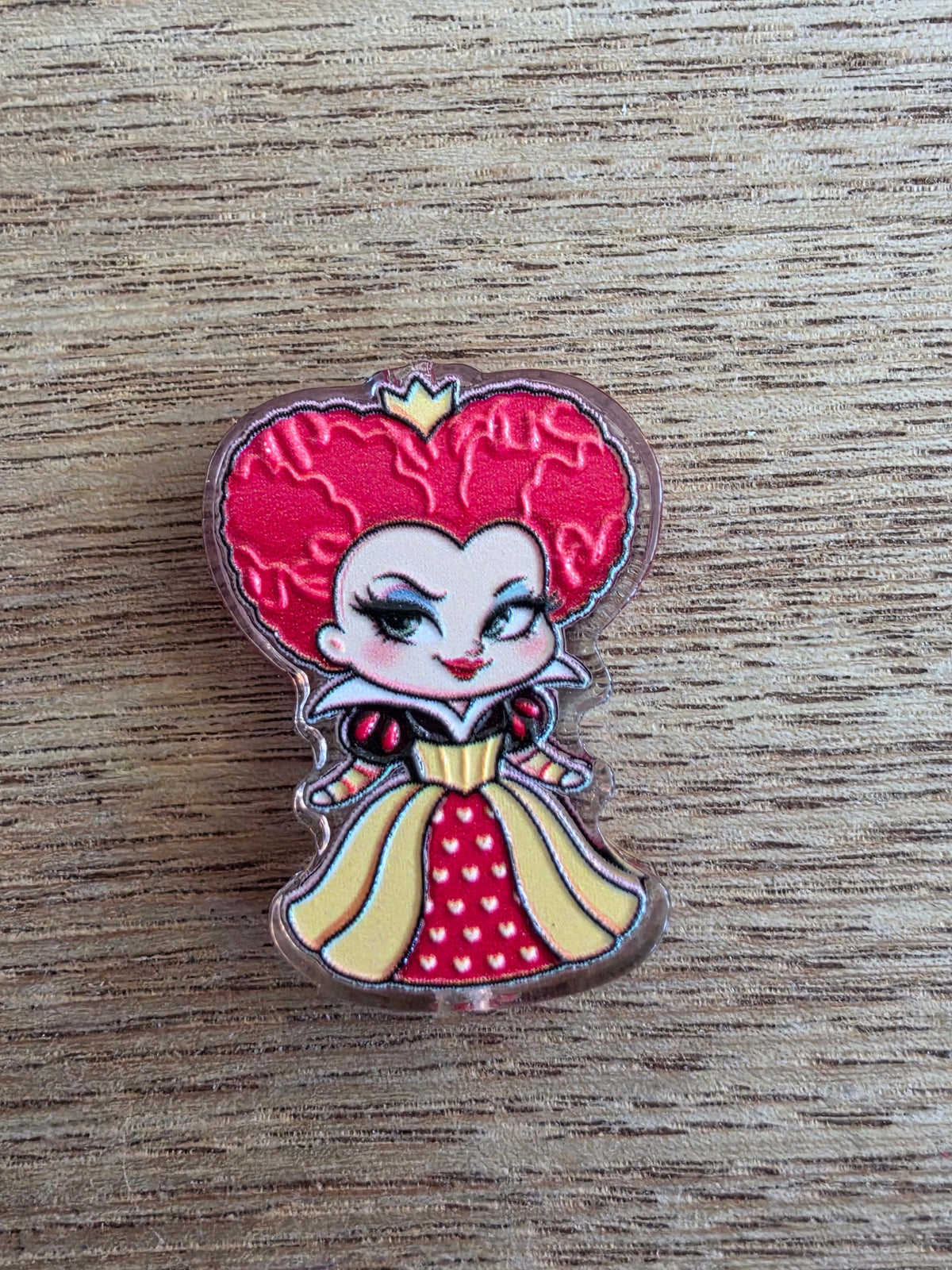 An acrylic bead shaped like a red heart, featuring a cartoon character with red hair, a red-and-white dress, and yellow accents, placed on a wooden surface.