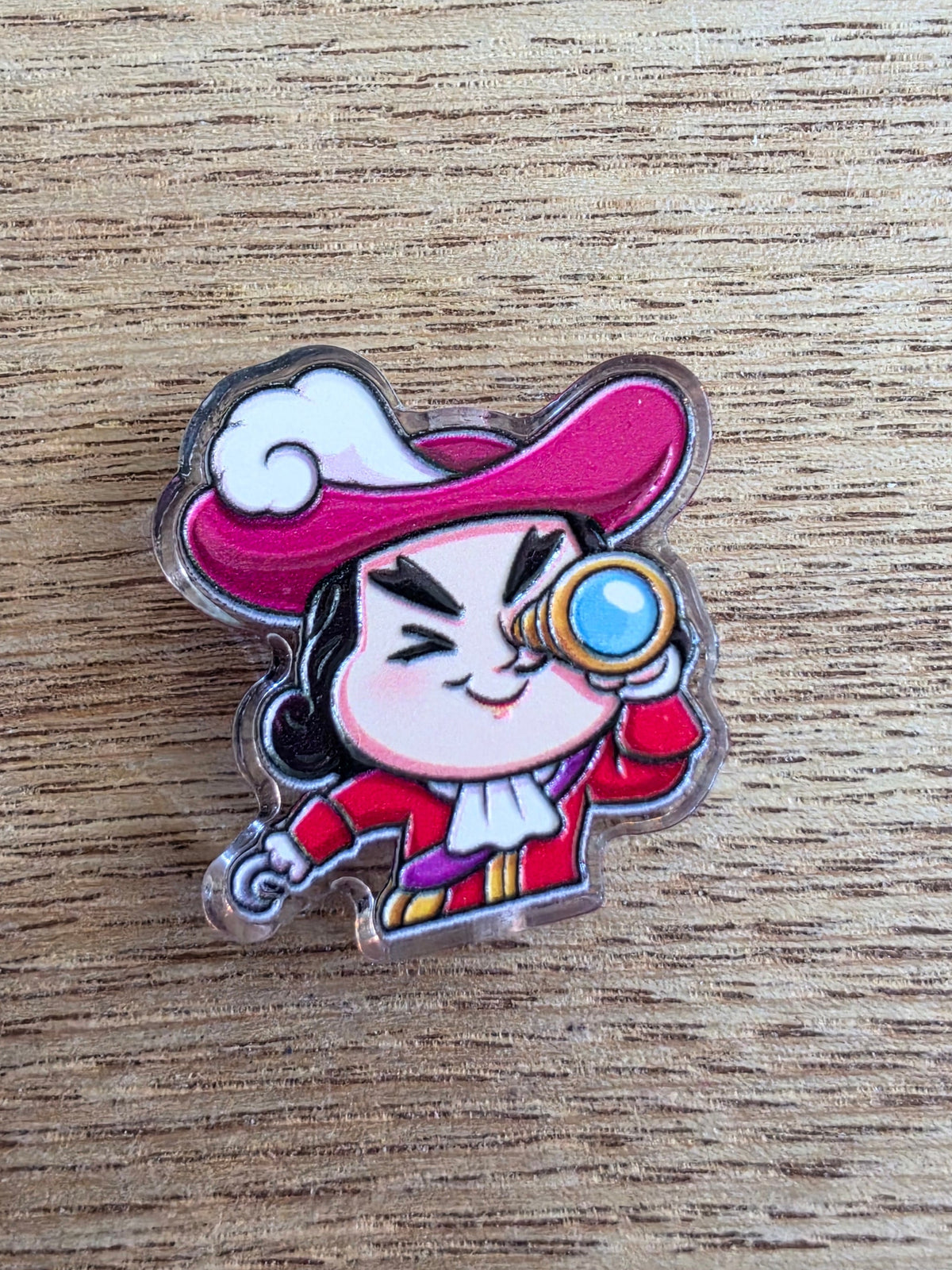 An acrylic bead featuring a cartoon pirate with a pink hat, red outfit, and blue eyepatch, photographed on a light wood surface.