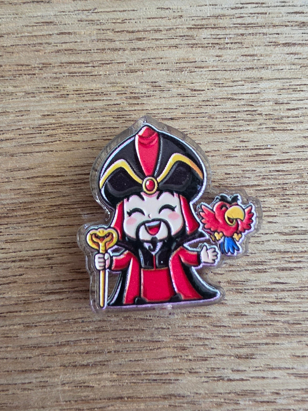 An acrylic bead shaped like a cartoon genie character in a red robe and black hat, holding a gold scepter and a red flower, placed on a light wood surface.
