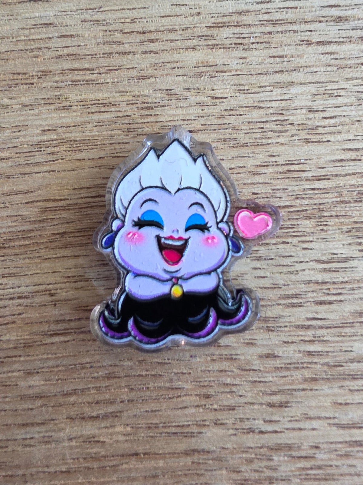 An acrylic bead of a cartoon character with white hair, blue eyes, and a pink heart, placed on a light brown wooden surface
