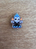 An acrylic bead featuring a cartoon chibi character with blue hair, a black outfit, and orange flames, placed on a light brown wood surface.