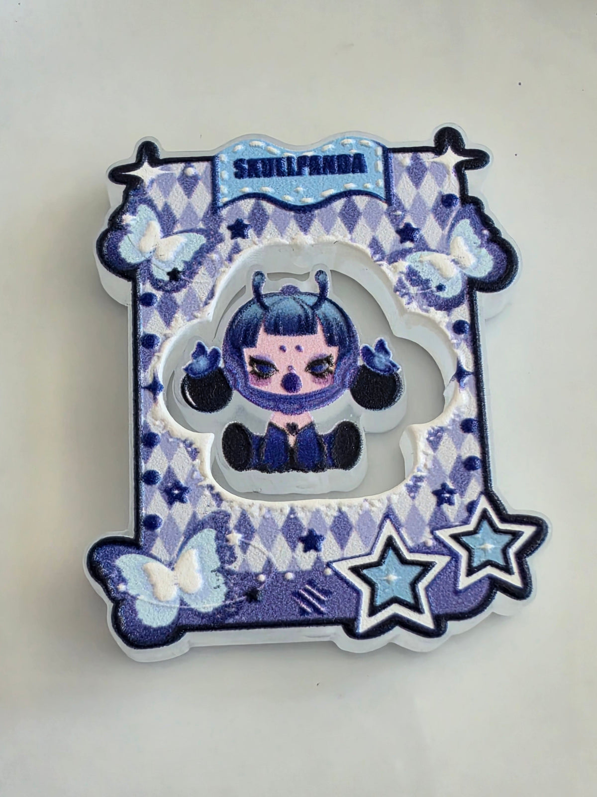 An acrylic bead set featuring a stylized cartoon character with dark blue hair and black horns, surrounded by a blue and white harlequin pattern, butterflies, and star details. The top banner reads 'SKULLPANDA'.
