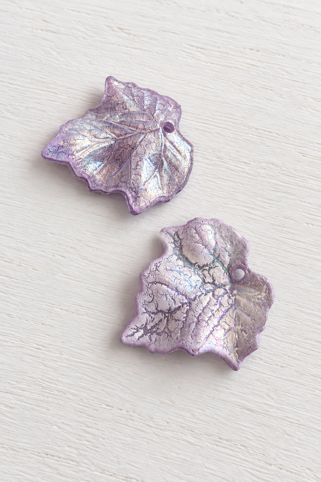 Two iridescent lavender-purple grapevine acetate leaves with a metallic sheen, displayed on a white wood background