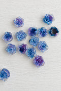 A group of small rose-shaped beads or charms in shades of blue, purple, and iridescent tones, scattered on a plain light background.