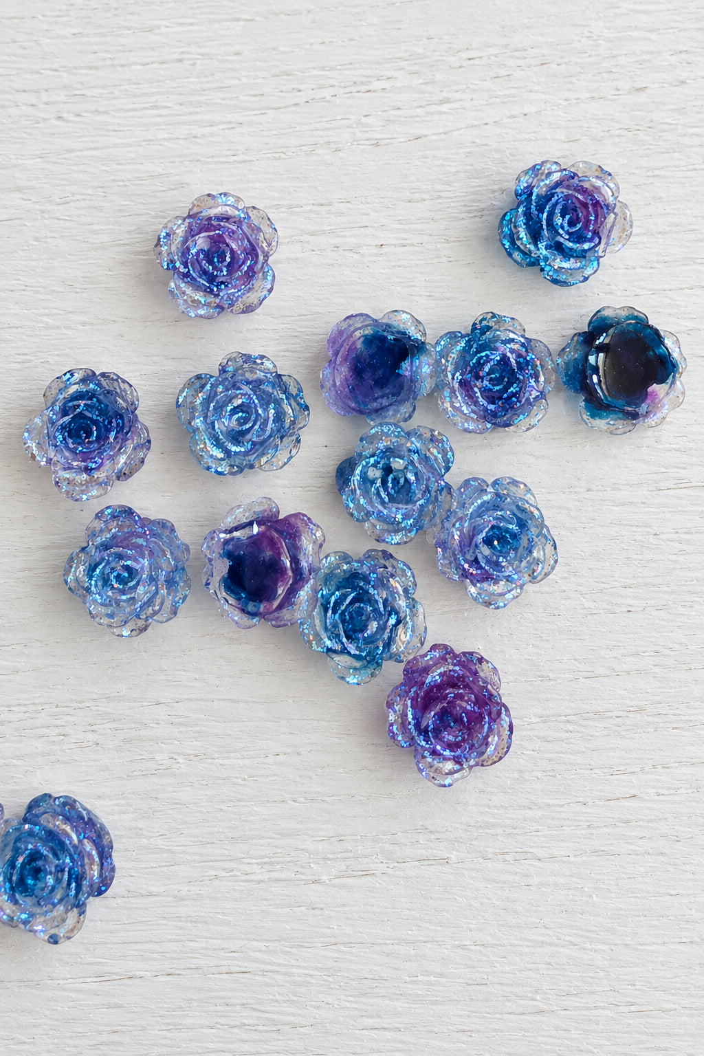 A group of small rose-shaped beads or charms in shades of blue, purple, and iridescent tones, scattered on a plain light background.