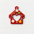 A small red decorative charm shaped like a house with a heart-shaped cut out in the centre, decorated with white flowers, a yellow star, and small colourful accents.