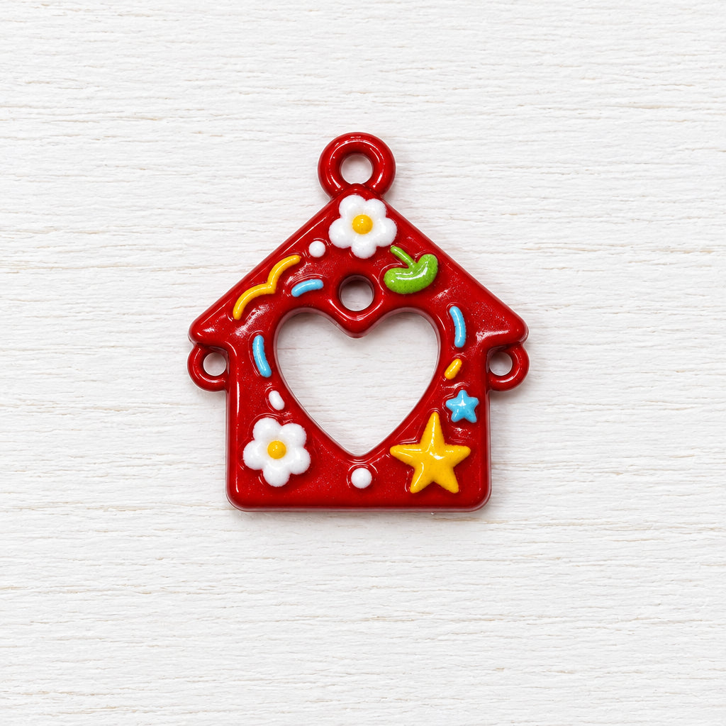 A small red decorative charm shaped like a house with a heart-shaped cut out in the centre, decorated with white flowers, a yellow star, and small colourful accents.