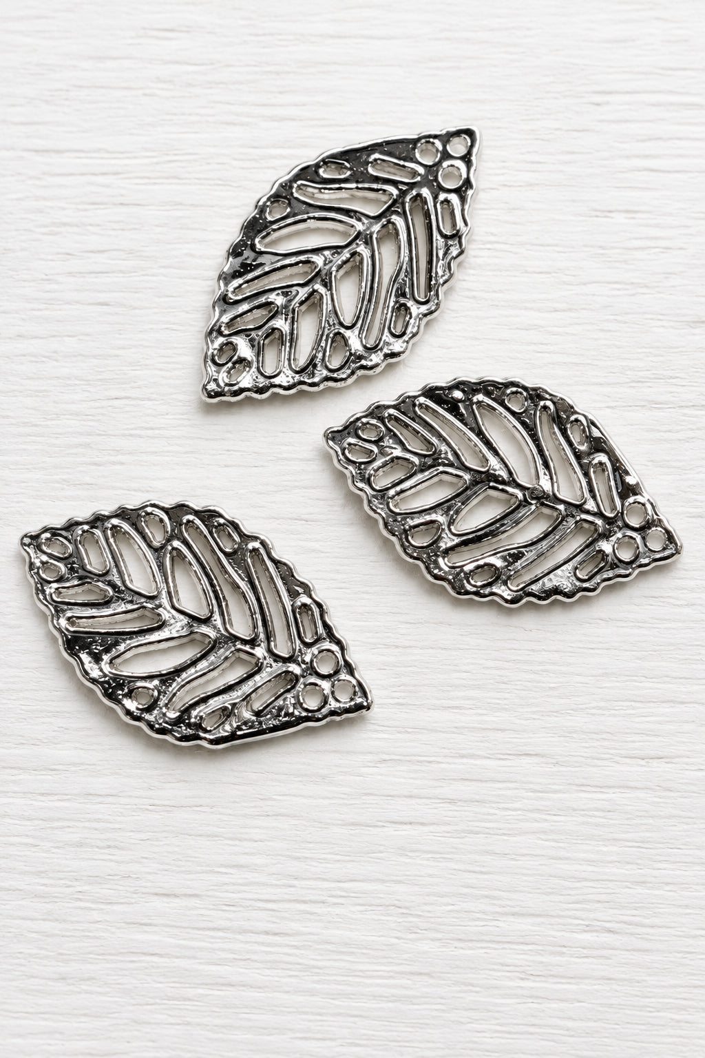 Acrylic silver leaf-shaped pendants with detailed cut out leaf patterns, displayed on a glossy white surface.