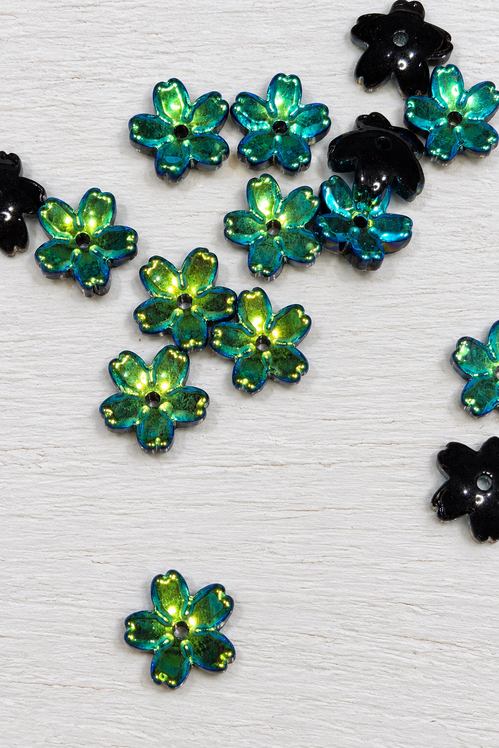 Small glass flower-shaped beads scattered on a white surface. The flowers are iridescent with a colour-shifting green and blue finish, with some pieces showing black edges or backing.