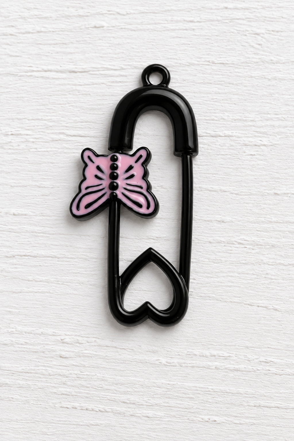 A black safety pin-shaped charm with a heart at the bottom and a pink butterfly accent on the side, shown on a plain white background.