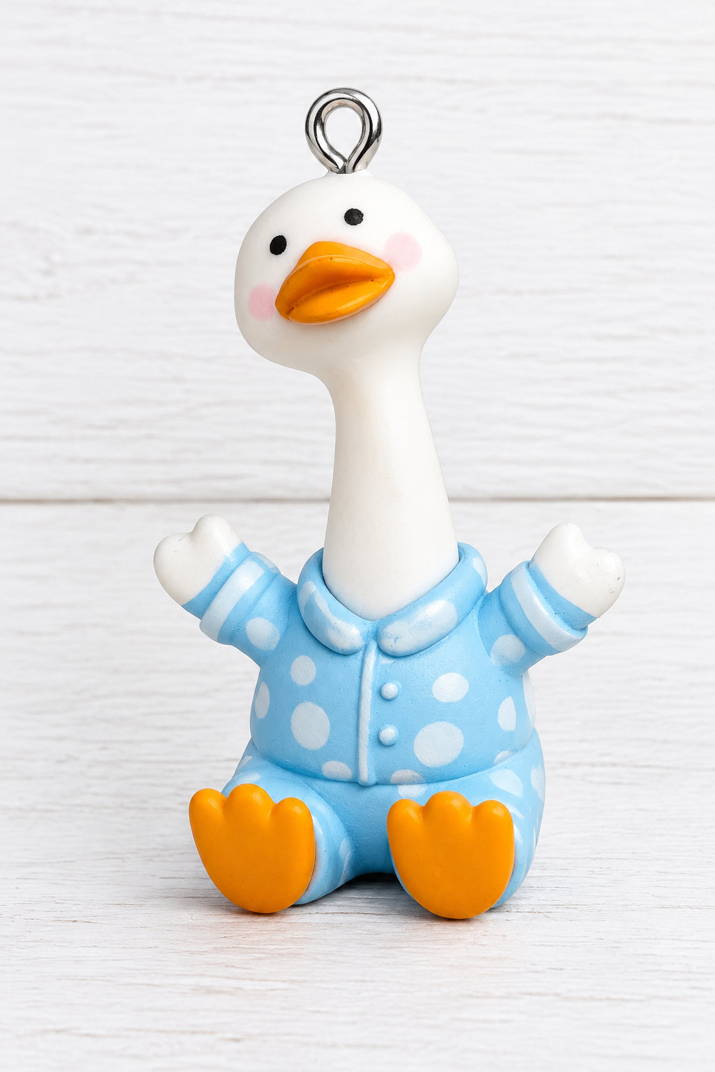 A small duck figurine wearing light blue pajamas with white polka dots, held in a person's hand. The duck has an orange beak, orange feet, and a white body, with a small metal loop at the top of its head.