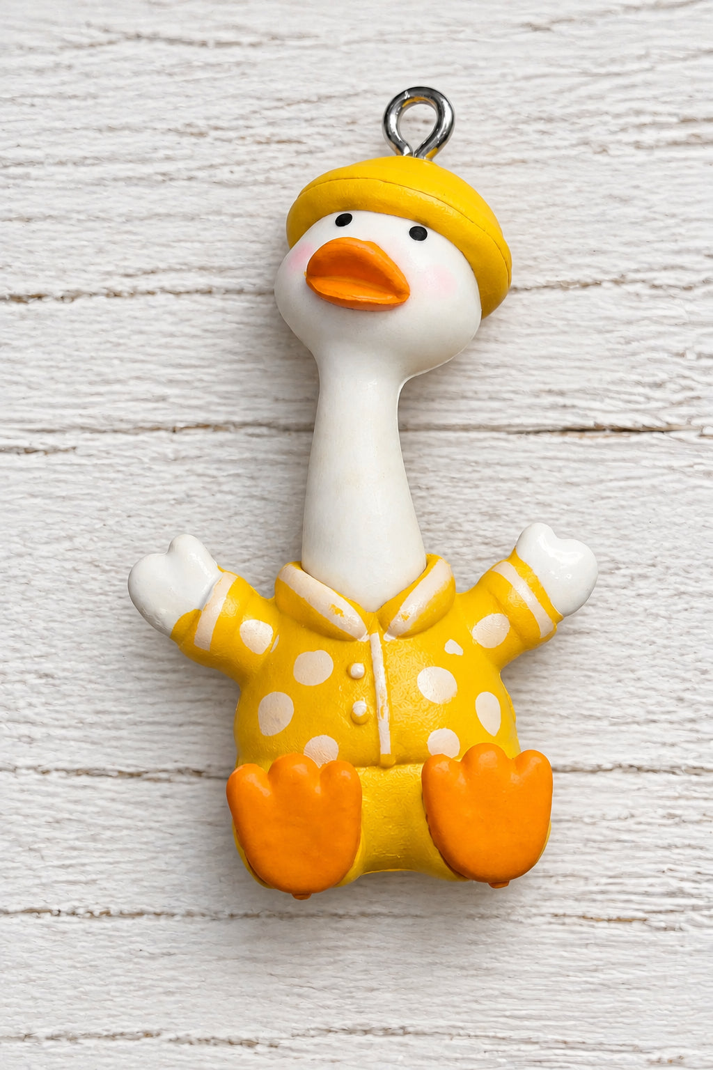 A small duck-shaped pendant. The duck has a white neck and face, orange beak and feet, and wears a yellow hat and yellow polka-dot outfit, with a metal loop at the top.