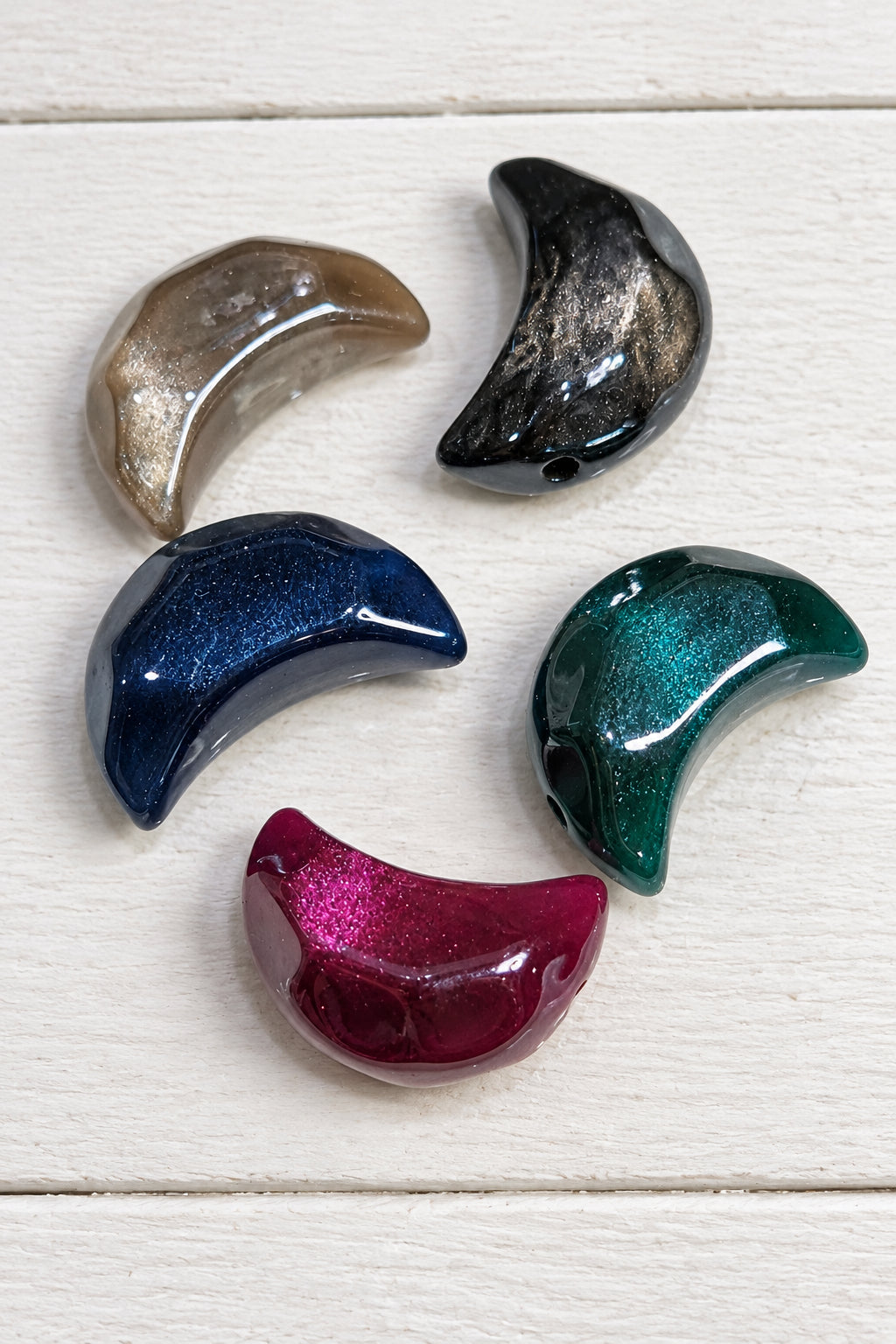 Five glossy crescent-shaped beads in assorted colours and finishes, including smoky brown, black, navy blue, emerald green, and magenta.