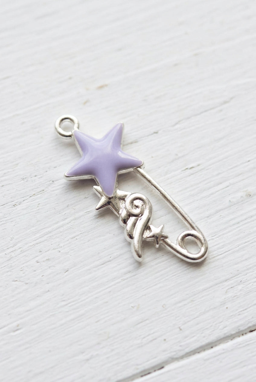 A silver safety pin charm featuring a lavender purple star on the top and a small silver crescent moon and star detail below, resting on a plain light surface.