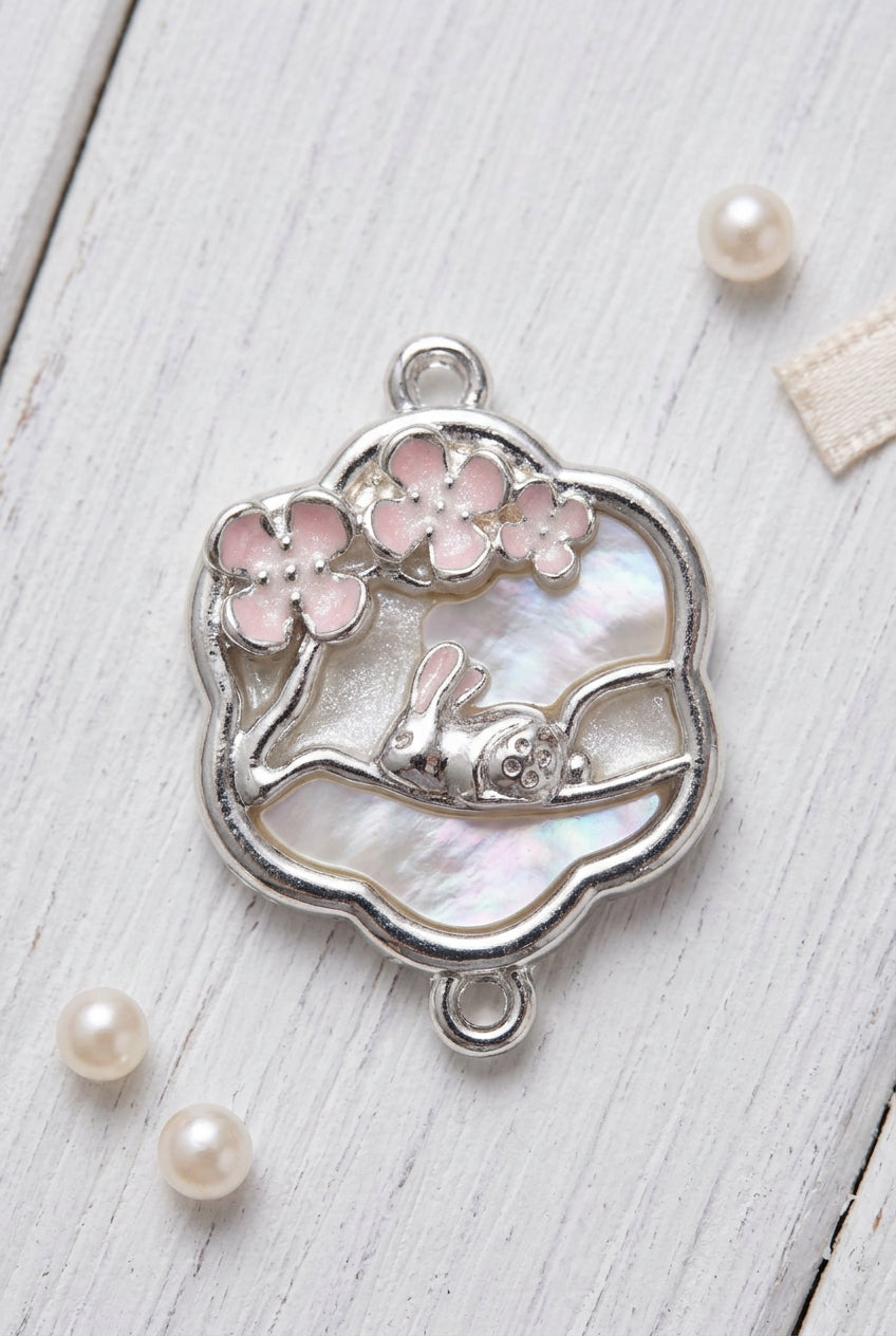A silver-toned metal charm featuring a sleeping bunny under pink cherry blossoms, displayed on a light gray surface.