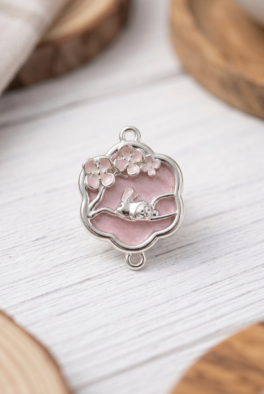 A silver-toned metal charm with a scalloped pink enamel border, featuring cherry blossom details and a small sleeping bunny figure, shown on a light gray surface.