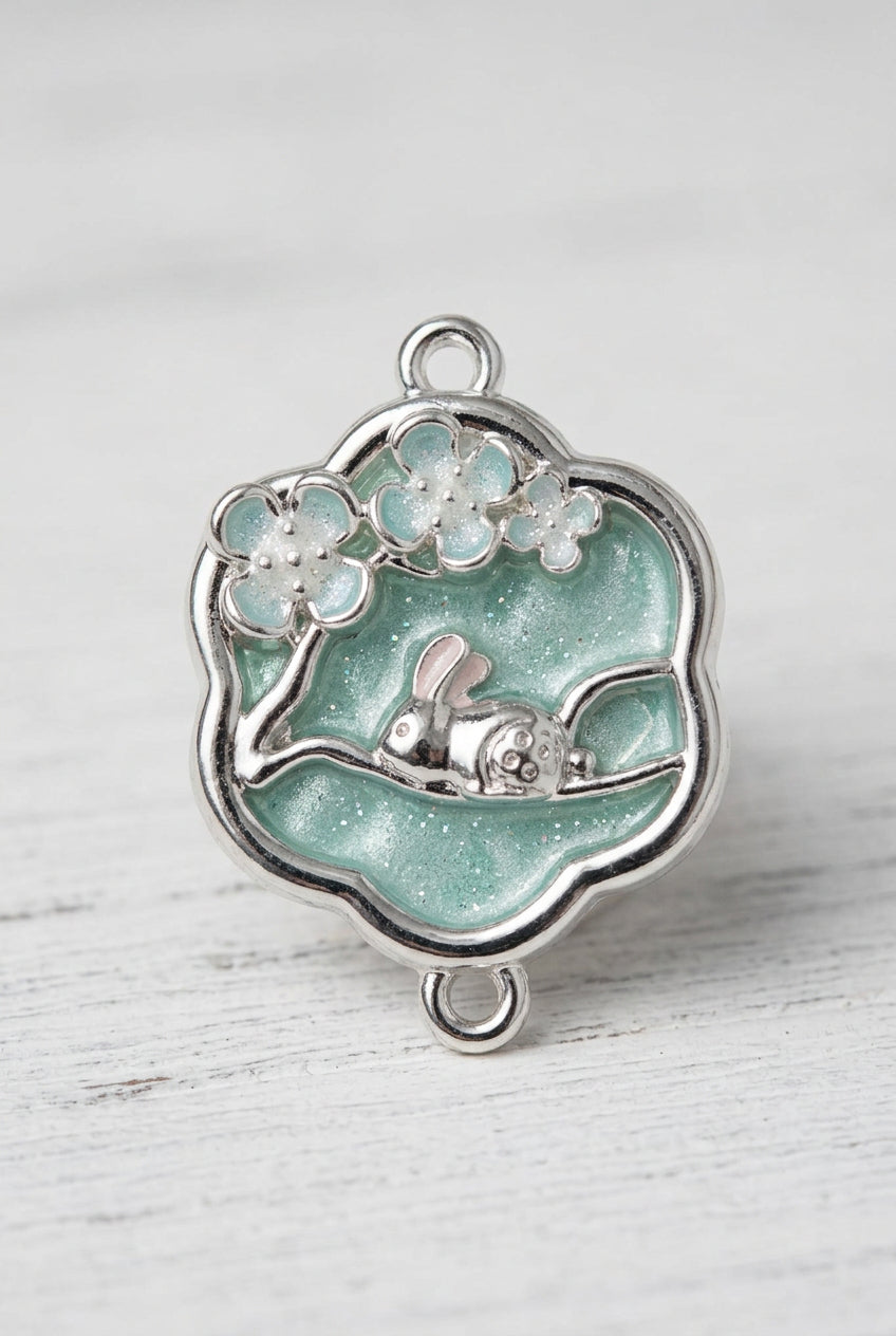 A silver-toned metal charm with a scalloped blue enamel border, featuring cherry blossom details and a small sleeping bunny figure, shown on a light gray surface.